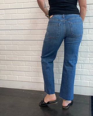 A woman wearing blue jeans and a black sleeveless top, photographed from behind against a white brick wall.