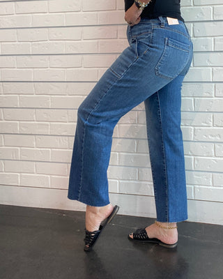A woman wearing blue straight-leg jeans, a black sleeveless top, and black sandals poses in a mirror selfie against a white brick wall.