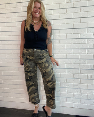 A woman wearing a black sleeveless top and camouflage cargo pants lies on her side against a white painted brick wall.