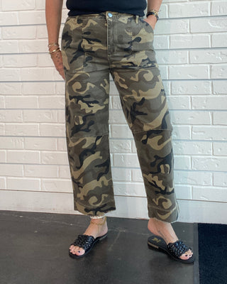A woman wearing camouflage cargo pants, a black sleeveless top, and black slide sandals, standing against a white brick wall.