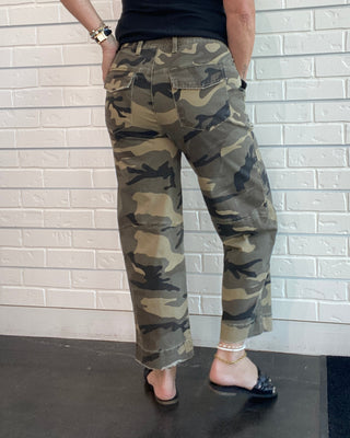 A person wearing camouflage cargo pants, a black tank top, and black flat shoes, standing against a white brick wall.