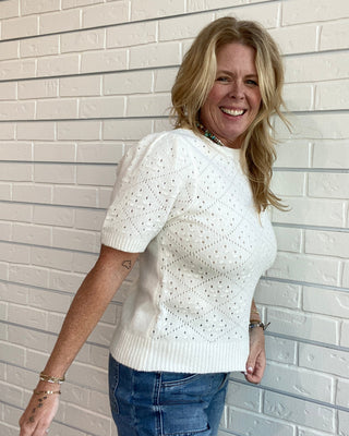 A smiling blonde woman wearing a white short-sleeve knit top with a textured eyelet pattern and blue jeans, standing against a white painted brick wall.