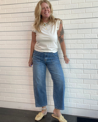 A woman stands against a white brick wall wearing a white sleeveless top, blue cropped jeans, and beige woven mule sandals.