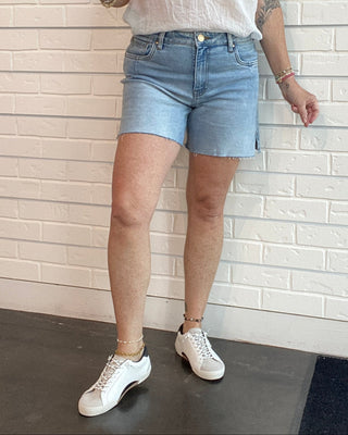 Woman wearing a white shirt and denim shorts standing against a white brick wall.