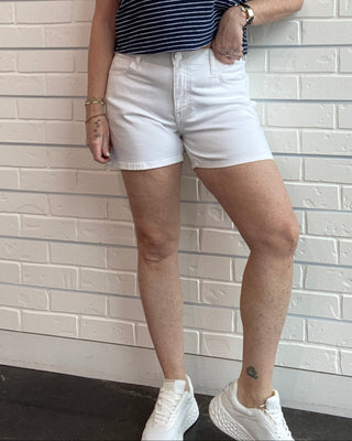 Woman wearing a striped tank top and white shorts standing against a light brick wall.