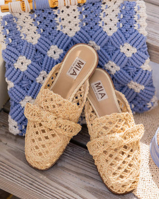 Pair of woven sandals with 'MIA' branding on a textured surface with a blue and white patterned bag in the background.