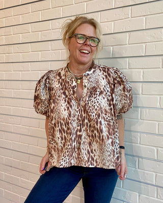 Woman wearing a leopard print blouse and blue pants standing against a beige brick wall.