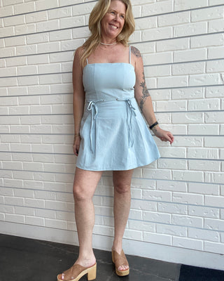 Woman wearing a light blue dress standing against a white brick wall.