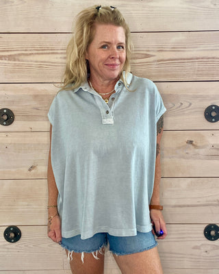Woman wearing a light blue sleeveless shirt and denim shorts standing against a wooden wall.