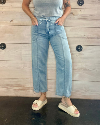 Person wearing a gray top and light blue jeans standing against a wooden wall.