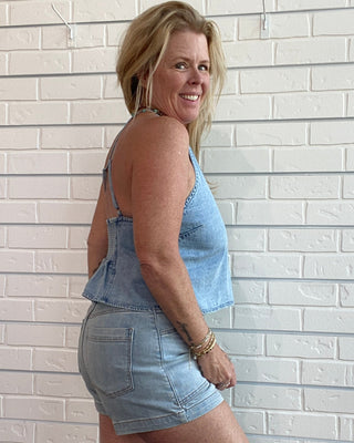 A woman posing in a light blue denim tank top and matching denim shorts against a white brick wall.