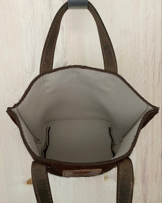 A brown leather tote bag shown from a top-down view, hanging from a gray hook against a light-colored wooden background.