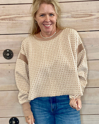 Woman wearing a beige sweater with brown patches and blue jeans standing against a wooden wall.