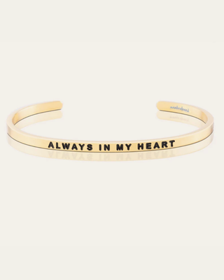 A gold-plated cuff bracelet with the phrase 'ALWAYS IN MY HEART' engraved in black capital letters on the front, displayed on a clean off-white background.