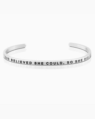 A silver metal cuff bracelet engraved with the motivational quote 'SHE BELIEVED SHE COULD, SO SHE DID' in black capital letters, shown on a clean white background.