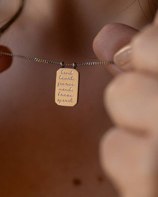 A close-up lifestyle shot of a person holding a delicate silver chain necklace with a small rectangular gold pendant engraved with the words 'kind hearts, peaceful mind, brave spirit'.