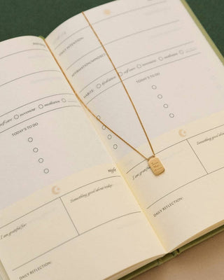 A gold necklace with a small engraved square pendant is laid across an open cream-colored planner or journal with lined daily reflection pages.