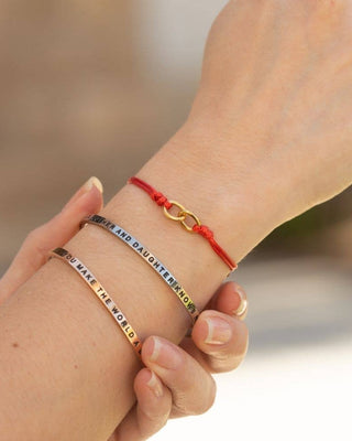 Three personalized bracelets worn on a person's wrist and hand, featuring a red cord bracelet with a gold infinity link, a silver chain with colorful lettering, and a gold bar engraved with 'YOU MAKE THE WORLD' in reverse.