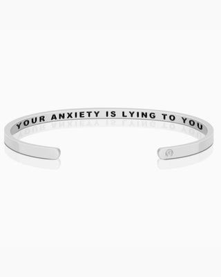 A silver open cuff bracelet engraved with the text 'YOUR ANXIETY IS LYING TO YOU' in black capital letters, shown on a clean white background with a soft reflection.