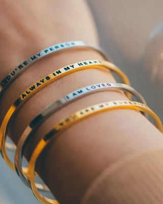 Four thin bangle bracelets stacked on a wrist, featuring silver and gold finishes with engraved romantic phrases like 'YOU'RE MY PERSON' and 'ALWAYS IN MY HEART'.