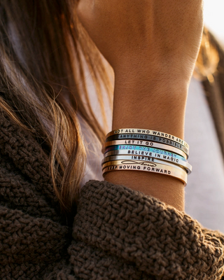 A close-up lifestyle shot of a person wearing a stack of five silver-toned metal bracelets on their wrist. The bracelets feature inspirational quotes engraved in black, blue, and silver lettering.