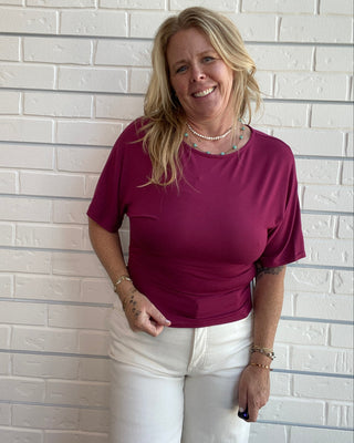 A smiling blonde woman wearing a short-sleeve magenta-purple t-shirt and white pants, standing against a white painted brick wall.