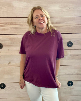Woman wearing a maroon shirt and white pants standing against a wooden wall.
