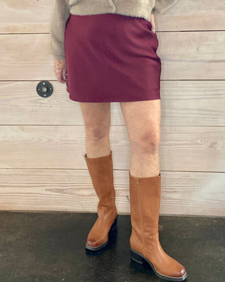 Woman wearing a beige sweater, maroon skirt, and brown knee-high boots against a wooden wall.