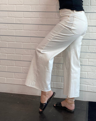 A woman wearing white wide-leg cropped pants, a black sleeveless top, and black strappy heels, lying on a white brick wall.