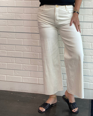 A woman modeling a pair of off-white cropped wide-leg pants with a black sleeveless top and black patterned slide sandals, standing against a white brick wall.
