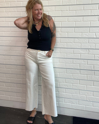 A woman wearing a black sleeveless top and white wide-leg pants, against a white brick wall.