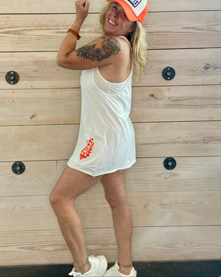 Person wearing a white tank top dress and orange cap against a wooden wall.