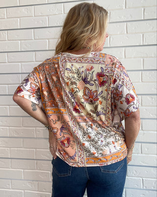 A woman viewed from behind wearing a short-sleeve bohemian-style top with a multicolor patchwork floral print, paired with blue jeans, standing against a white brick wall.