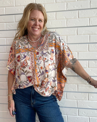 A woman with blonde hair smiling while wearing a bohemian-style short-sleeve blouse with a multicolor paisley and floral print, paired with blue jeans, standing against a white brick wall.