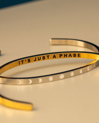 A close-up of three slim metallic cuff bracelets arranged on a light beige surface. One bracelet is gold-toned with the black engraved text 'IT'S JUST A PHASE', while the other two are silver-toned with white moon-phase dot details.