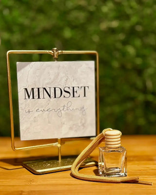 Small clear bottle with a yellow cap on a wooden surface next to a gold stand with a white card displaying 'MINDSET is everything'.