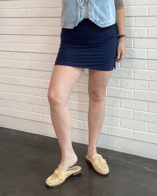 Woman wearing a denim vest over a black top and skirt against a white brick wall.