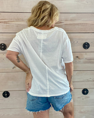 Person wearing a sheer white top over a plain shirt with denim shorts, standing against a wooden wall.
