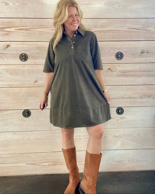 Woman wearing a green dress and brown boots standing against a wooden wall.