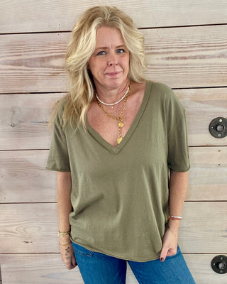 Woman wearing an olive green shirt and blue jeans standing against a wooden wall.