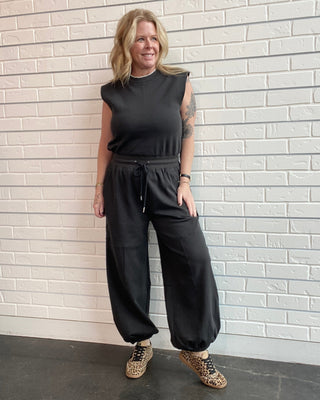 Woman wearing a black sleeveless top and pants standing against a white brick wall.