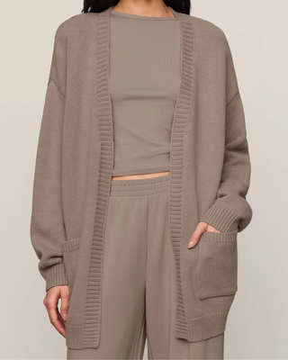 A woman modeling a taupe knit cardigan layered over a matching ribbed crop top and wide-leg pants against a plain light beige studio background.