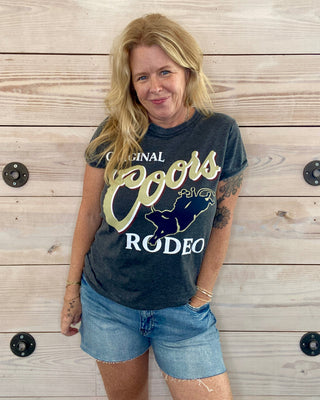 Woman wearing a Coors Rodeo t-shirt standing against a wooden wall.