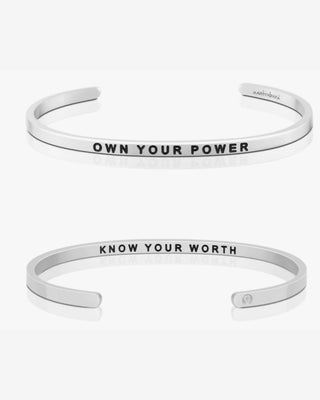 Two silver open cuff bracelets on a light gray background. The top bracelet is engraved with the words 'OWN YOUR POWER' and the bottom bracelet is engraved with 'KNOW YOUR WORTH'.