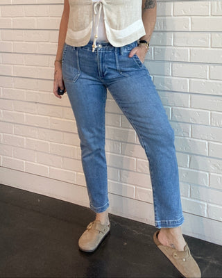 Woman standing against a light brick wall wearing a beige vest, white top, blue jeans, and beige shoes.