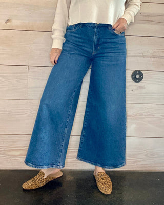 Woman wearing a white sweater and blue jeans standing against a wooden wall.