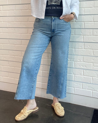 Woman wearing a white jacket, blue jeans, and a graphic t-shirt against a white brick wall.
