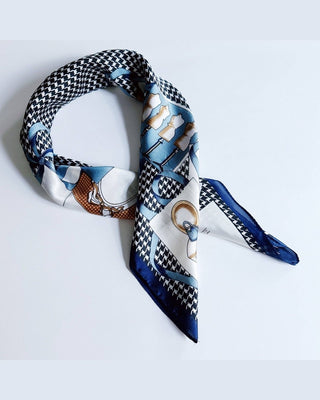 A square silk scarf with a complex graphic print featuring a black and white houndstooth pattern, blue and white stripes, and a brown-and-white checkered section, styled in a loop on a light gray background.