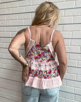 A woman viewed from behind wearing a tiered spaghetti-strap tank top with a colorful floral print and light pink lace trim, paired with light-wash jeans, standing against a white brick wall.