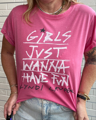 A woman wearing a pink graphic t-shirt with white text that reads 'GIRLS JUST WANNA HAVE FUN' and 'CLYNDI LIPPO' printed on the front, paired with light-wash denim jeans.